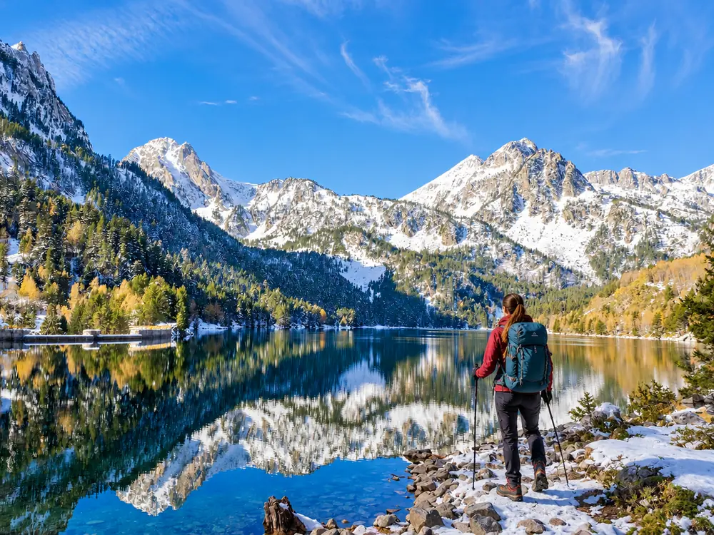 Senderista con mochila y bastones junto a un lago de montaña que refleja cumbres nevadas y bosque de otoño
