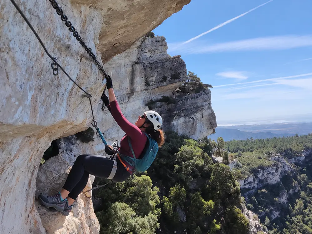 Climber on a via ferrata wearing a helmet and harness, clipped to the chain and cable on a rock wall above a forest